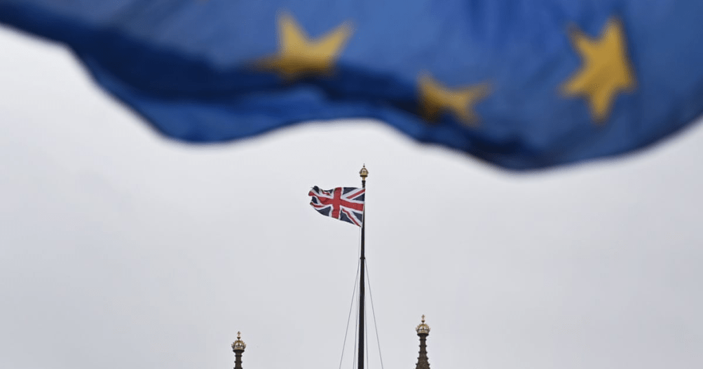 The image features a large, in-focus European Union (EU) flag billowing in the foreground, with the British Union Jack flag and the spires of the Palace of Westminster (including the Elizabeth Tower, popularly known as Big Ben) visible in the background. (Photograph: Andy Rain/EPA)